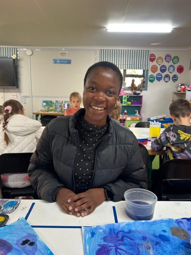 Vrouw in een klaslokaal, lachend en zittend achter een tafel met schilderkunstmateriaal.